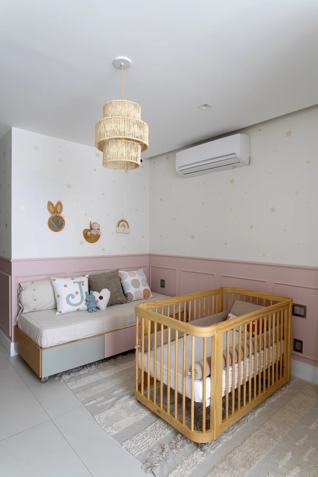 Quarto de bebê com berço de madeira, meia-parede rosa e luminária de fibra natural