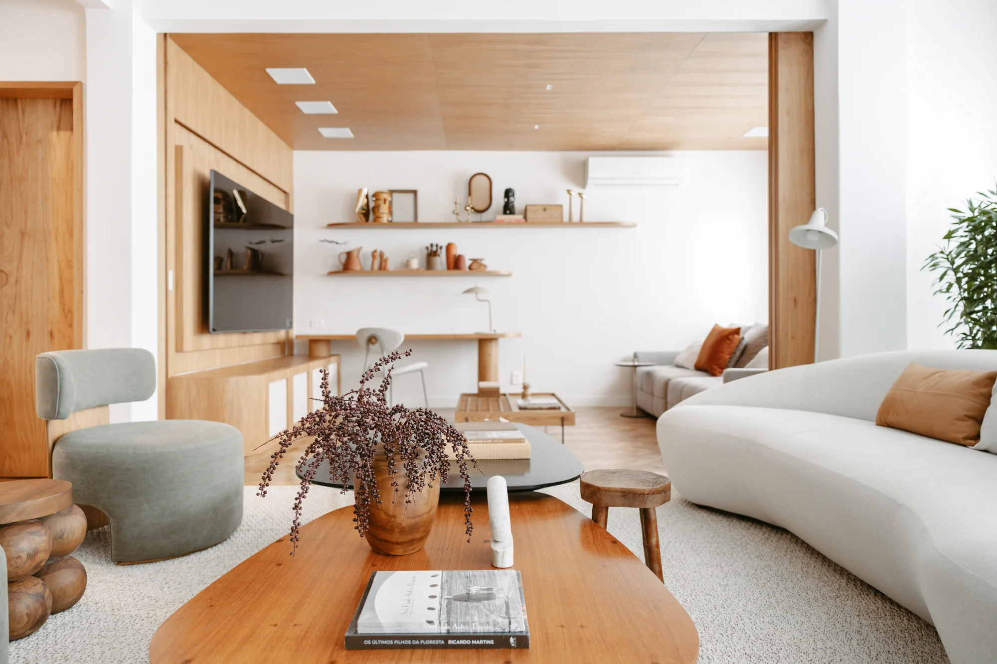 Living visto da mesa de centro, com painel de madeira e sofá curvo ao fundo