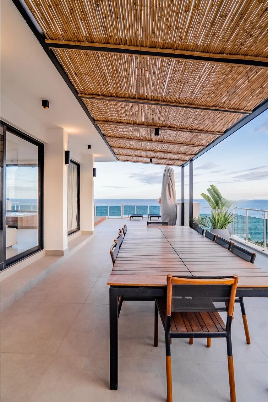 Varanda comprida com forro de bambu, mesa de jantar externa e vista para o mar