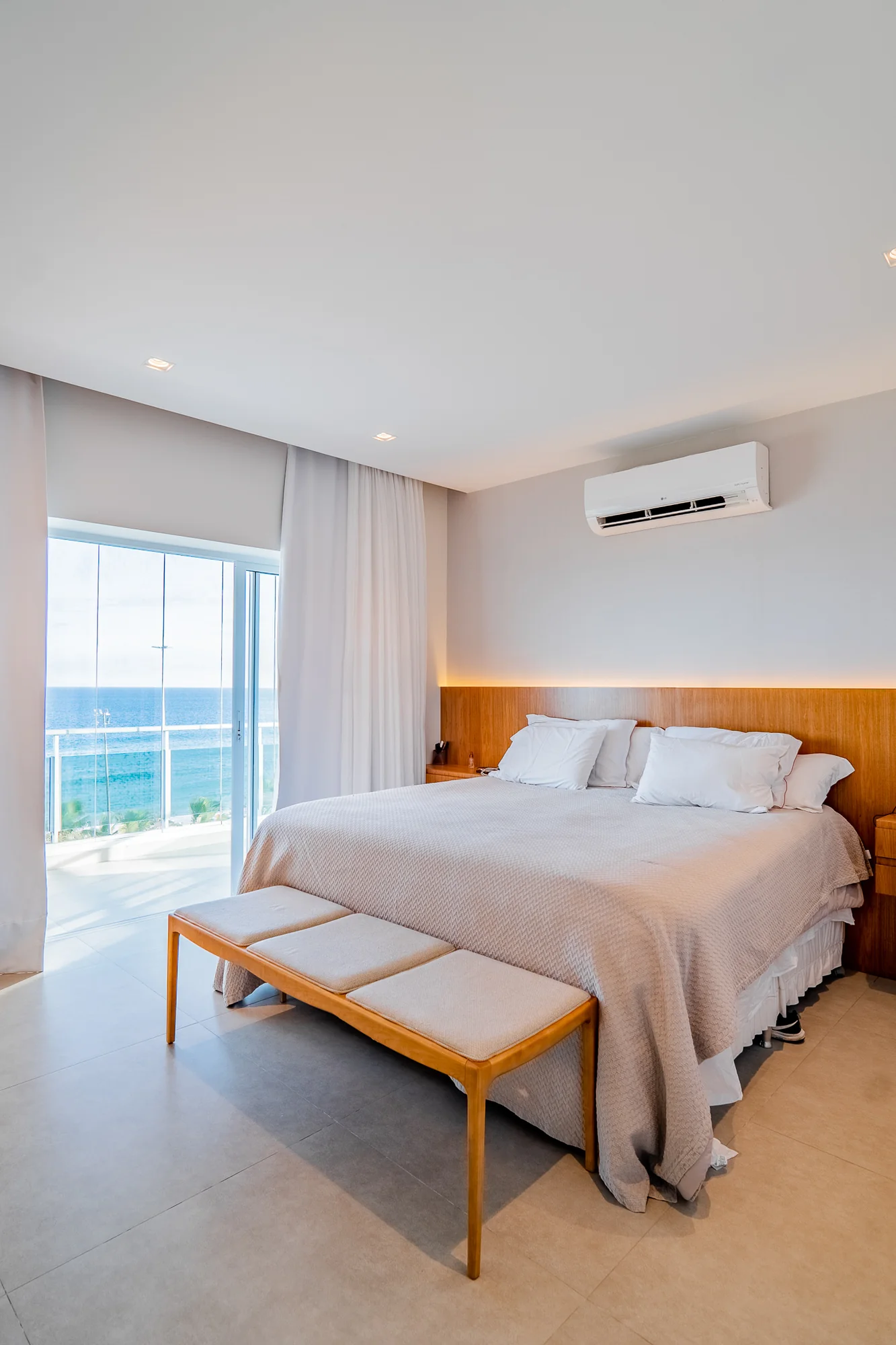 Quarto de casal com cabeceira de madeira, recamier ao pé da cama e janela para o mar